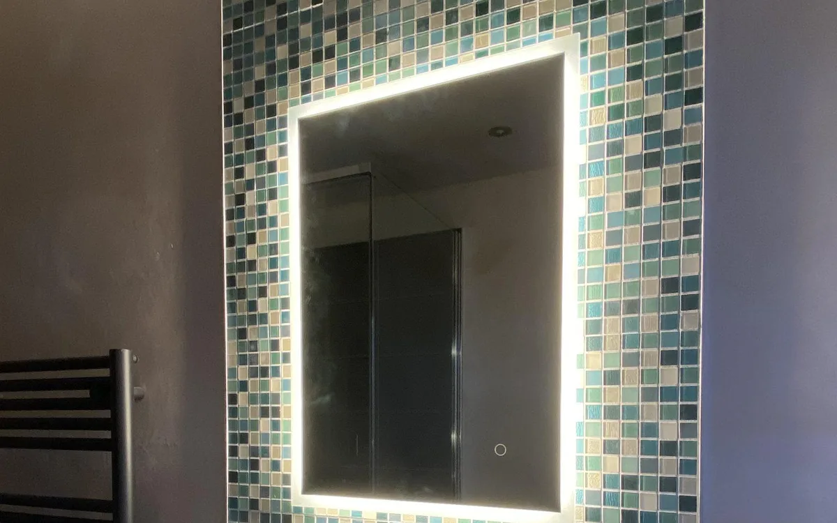 Custom LED backlit bathroom vanity mirror above a white sink with mosaic tile wall