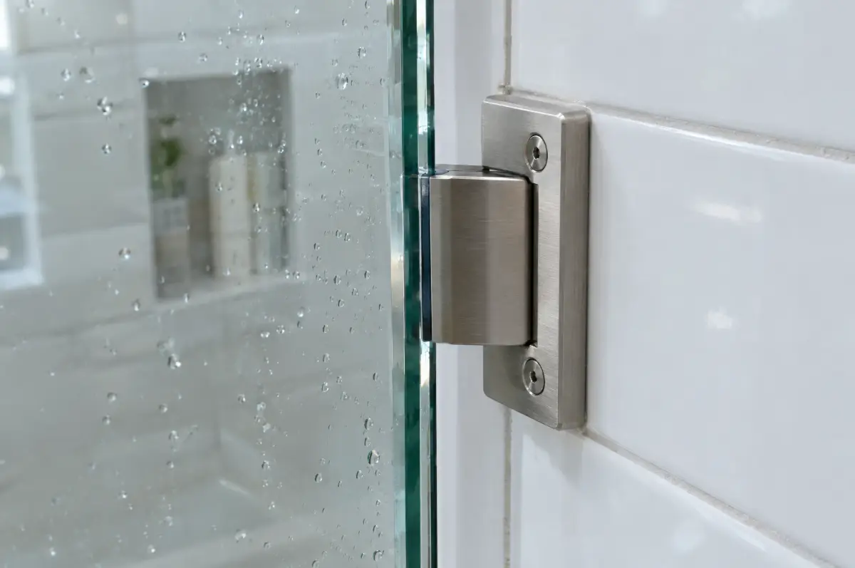Close-up of a brushed nickel hinge holding a thick frameless glass shower door, with the small gap between the glass and the tiled wall